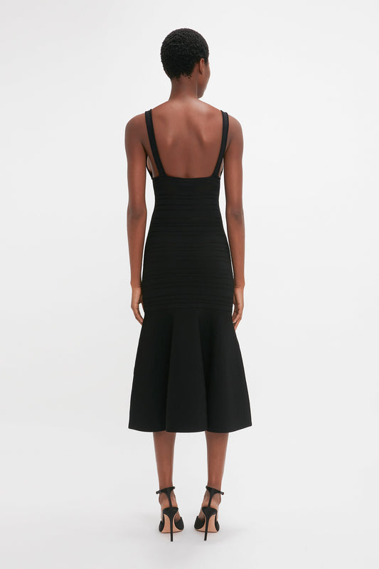 A woman from behind wearing a Victoria Beckham Frame Detail Sleeveless Dress in Black with a fit-and-flare silhouette and flared skirt, paired with black high heels, standing against a white background.