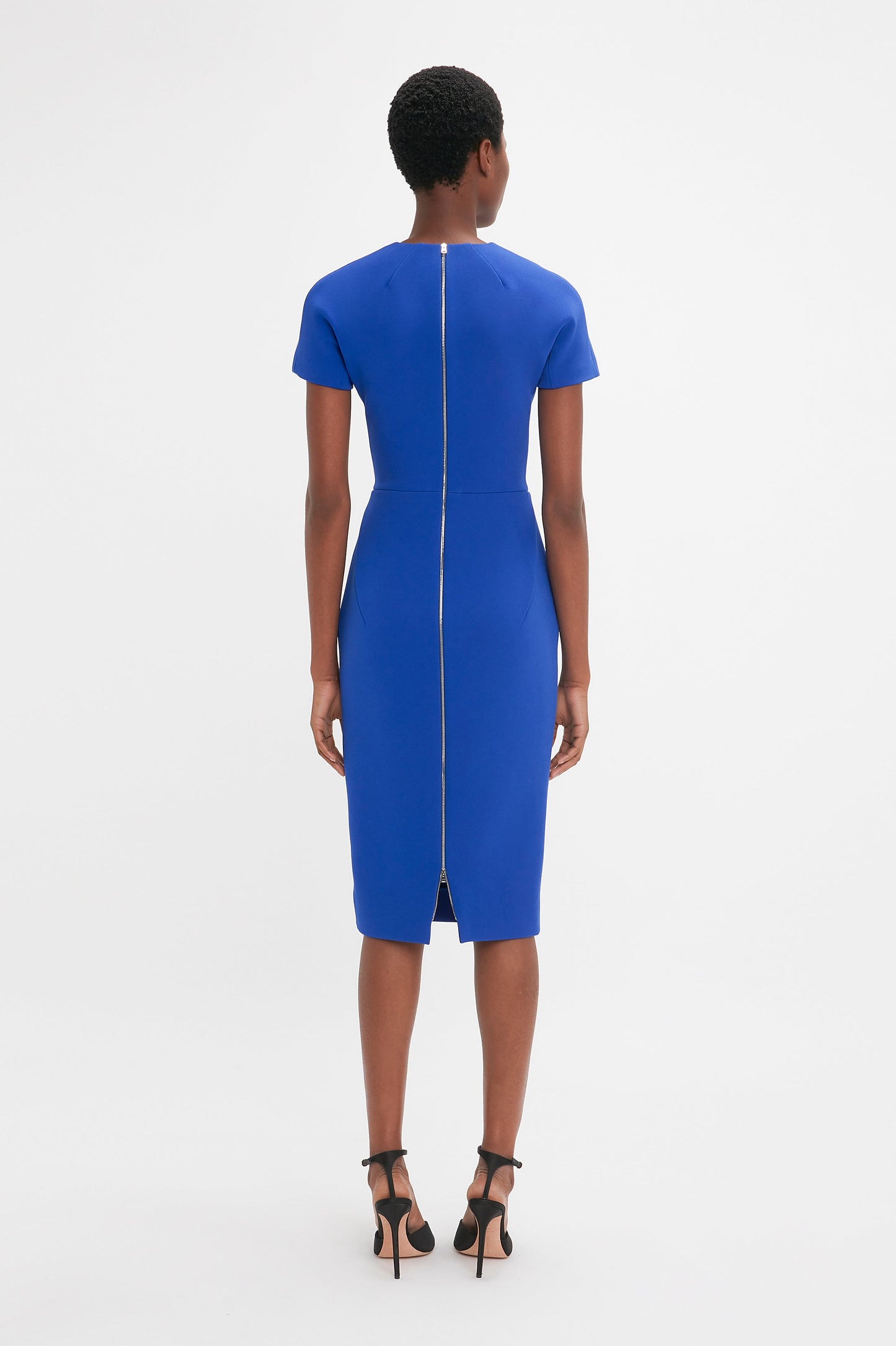 A woman stands facing away, wearing a Victoria Beckham fitted t-shirt dress in palace blue with a back zipper, paired with black heels against a white background.