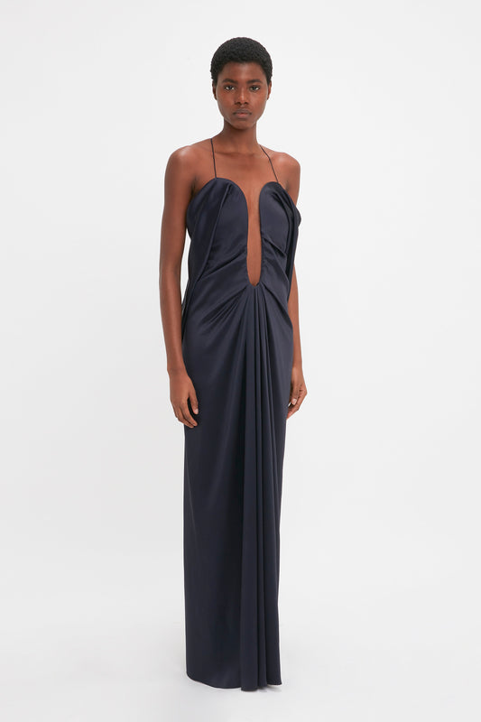 A young black woman models a strapless, navy blue crepe back satin evening gown with a front cutout, standing against a white background by Victoria Beckham.
