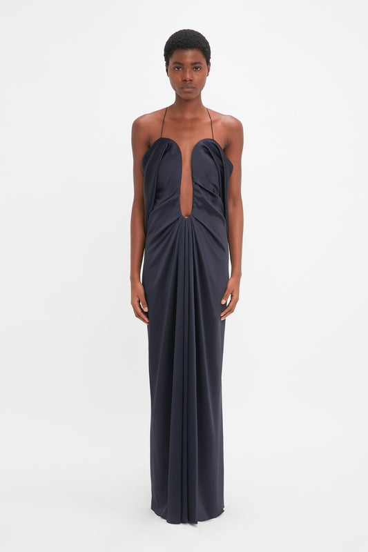 A black woman stands against a white background, wearing a long, elegant Victoria Beckham crepe back satin evening gown with thin straps and a keyhole neckline.