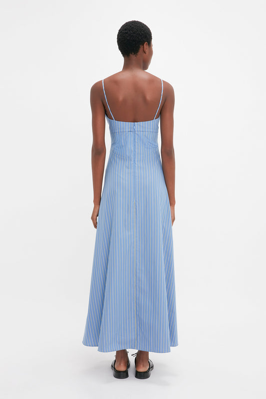 A woman viewed from behind, wearing a Victoria Beckham blue and white striped cotton sleeveless maxi dress and black heels, standing against a plain white background.