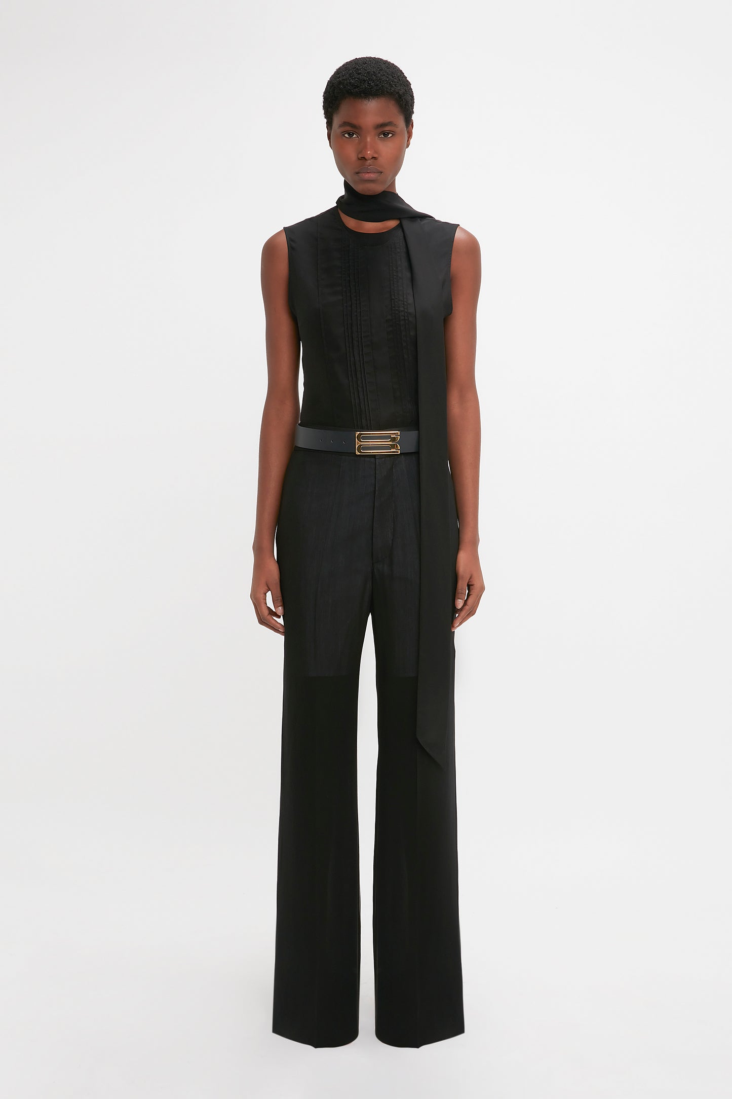 A black man models a modern black sleeveless top with a high collar and Victoria Beckham's Waistband Detail Straight Leg Trouser In Black in a white studio setting.