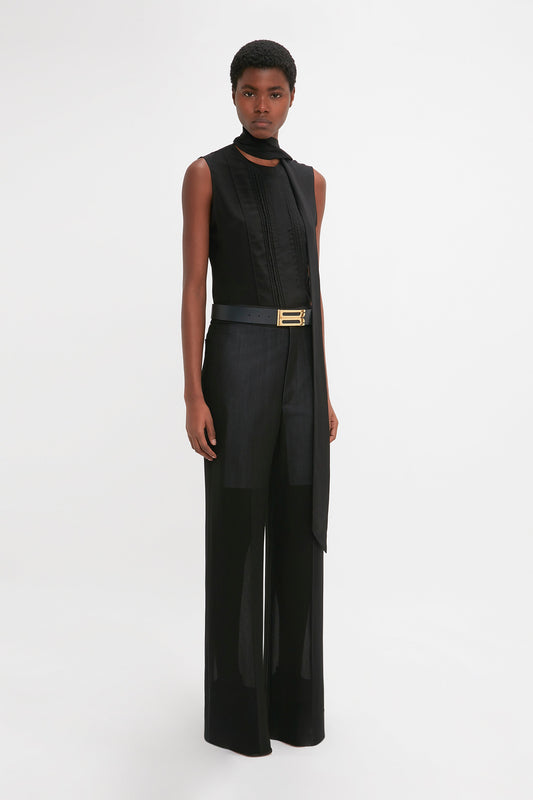 A young woman wearing Victoria Beckham's Waistband Detail Straight Leg Trouser in black with a high collar and sleek straight leg trousers, accessorized with a gold belt, stands against a plain white background.
