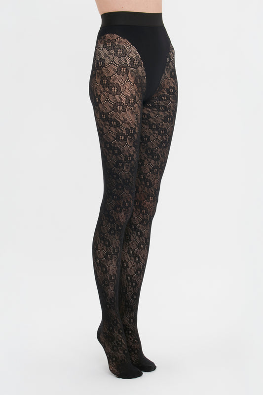 A person wearing black Victoria Beckham Monogram Lace Tights stands against a white background, side view.