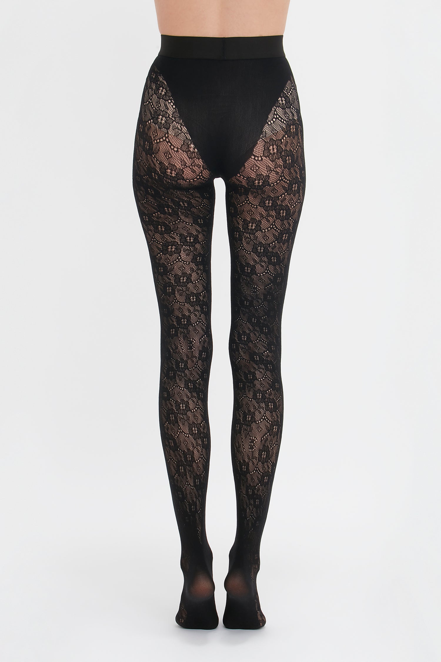 A person stands facing away from the camera, modeling Victoria Beckham's Exclusive VB Monogram Lace Tights In Black with seamless and sag-free construction against a white background.