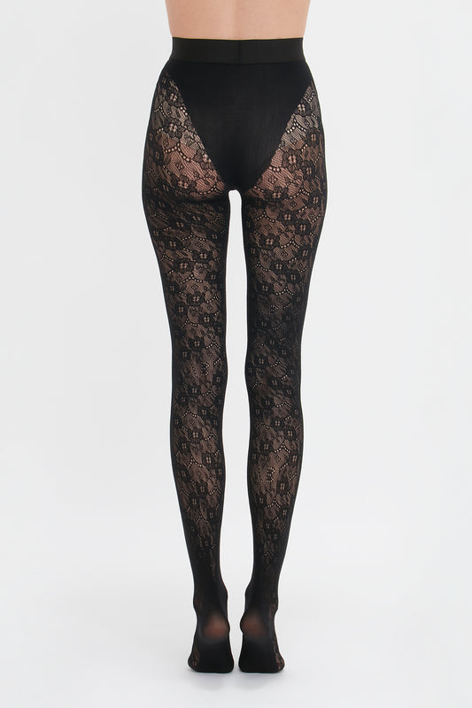 A person stands facing away from the camera, modeling Victoria Beckham's Exclusive VB Monogram Lace Tights In Black with seamless and sag-free construction against a white background.