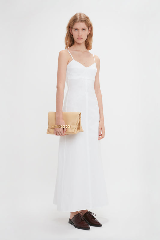 A woman in a white dress stands holding a Victoria Beckham Chain Pouch With Strap In Sesame Leather, with brown shoes placed in front.