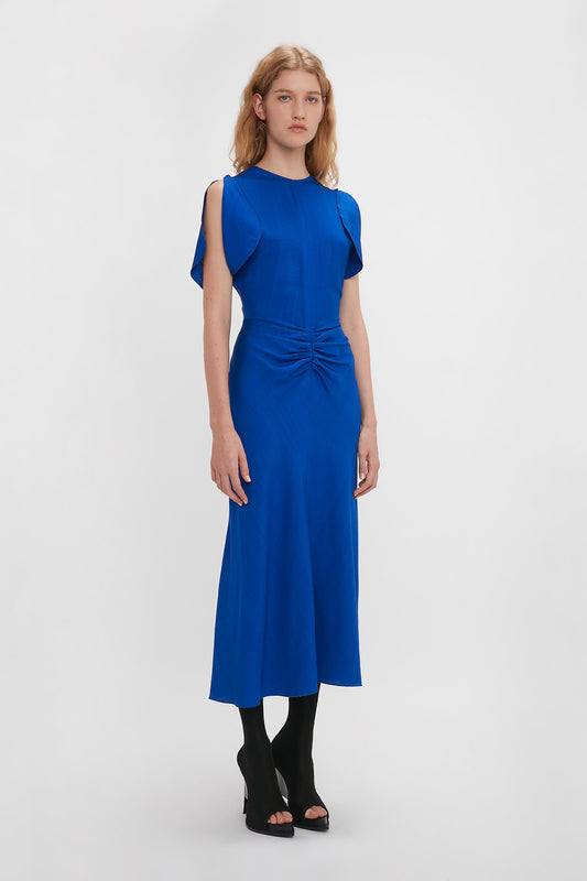 A woman in a Victoria Beckham palace blue gathered waist midi dress with cap sleeves and a fit-and-flare silhouette, paired with black heeled sandals, against a white background.