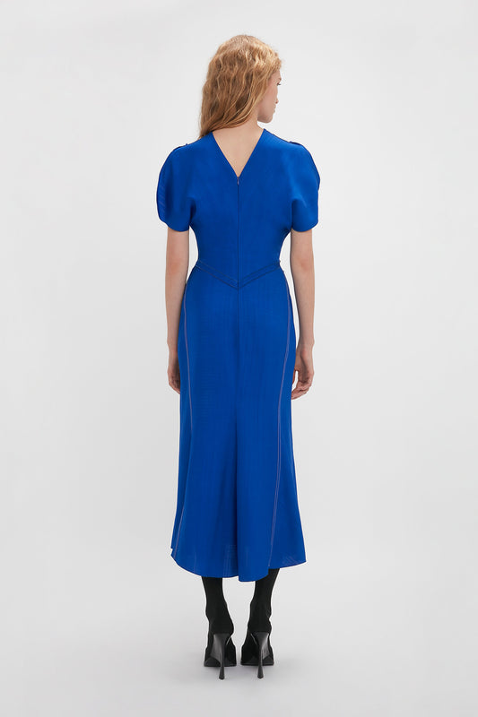 A woman in a Victoria Beckham Gathered Waist Midi Dress In Palace Blue and black heels stands with her back to the camera, emphasizing the dress's v-shaped neckline and pleated skirt.