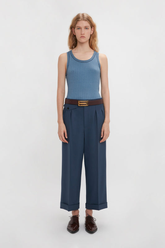 A young woman stands against a white background wearing a blue fine knit tank top, Victoria Beckham's Wide Leg Cropped Trouser In Heritage Blue, and brown leather shoes.