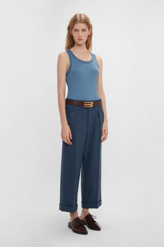 A woman in a blue fine knit tank and Victoria Beckham wide-leg cropped navy trousers with a brown belt, standing against a plain white background.