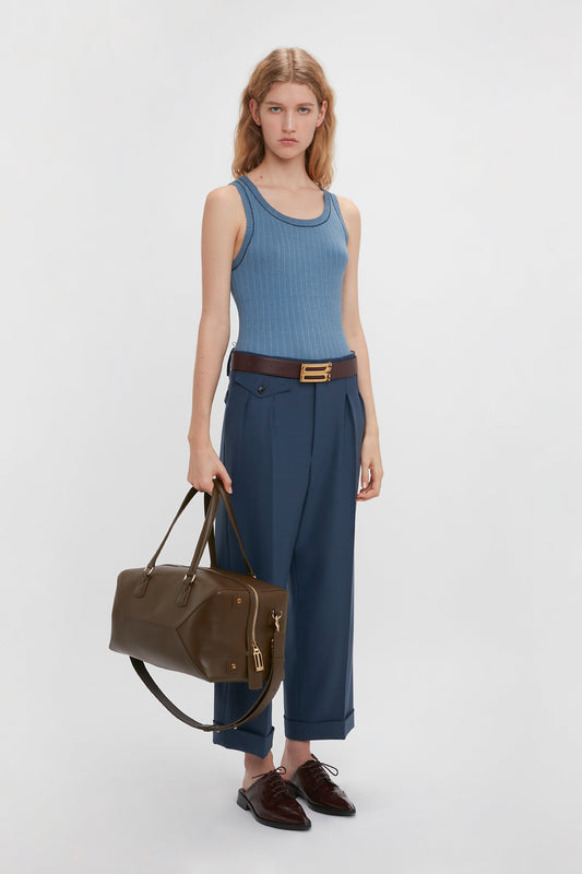 A woman in a fine knit tank top and Victoria Beckham wide leg cropped trousers in heritage blue, holding a brown leather bag, stands against a white background.