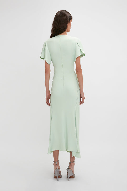 A woman from behind wearing a Victoria Beckham Gathered V-Neck Midi Dress In Jade with short sleeves and heels, standing against a plain background.