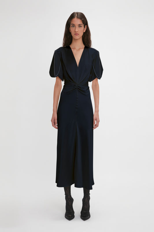 A woman in a sophisticated navy blue Exclusive Gathered V-Neck Midi Dress In Navy with puff sleeves, paired with black lace-up boots and VB Monogram Lace Tights, standing against a plain background by Victoria Beckham.