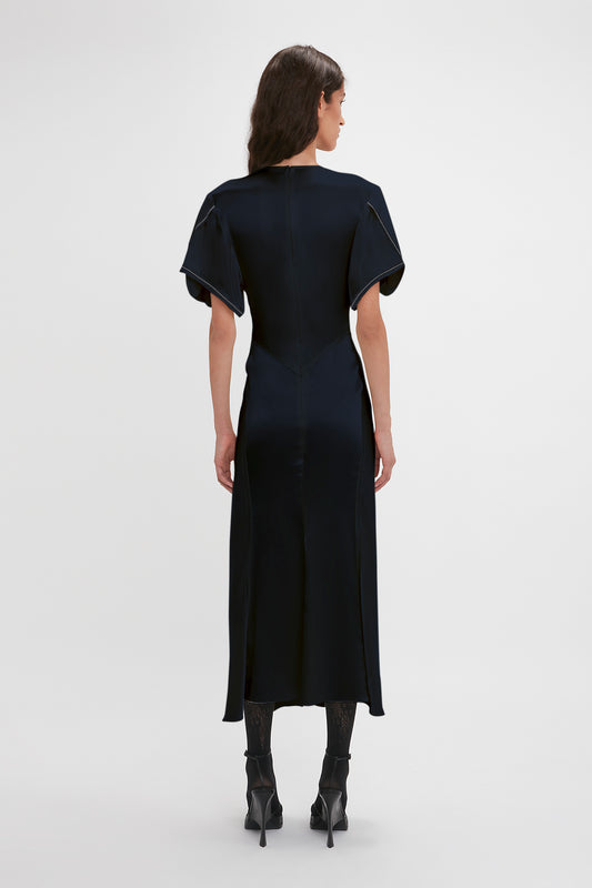 A woman stands facing away, wearing a navy blue Exclusive Gathered V-Neck Midi Dress by Victoria Beckham with short sleeves and a mid-calf hemline, paired with black heeled boots.