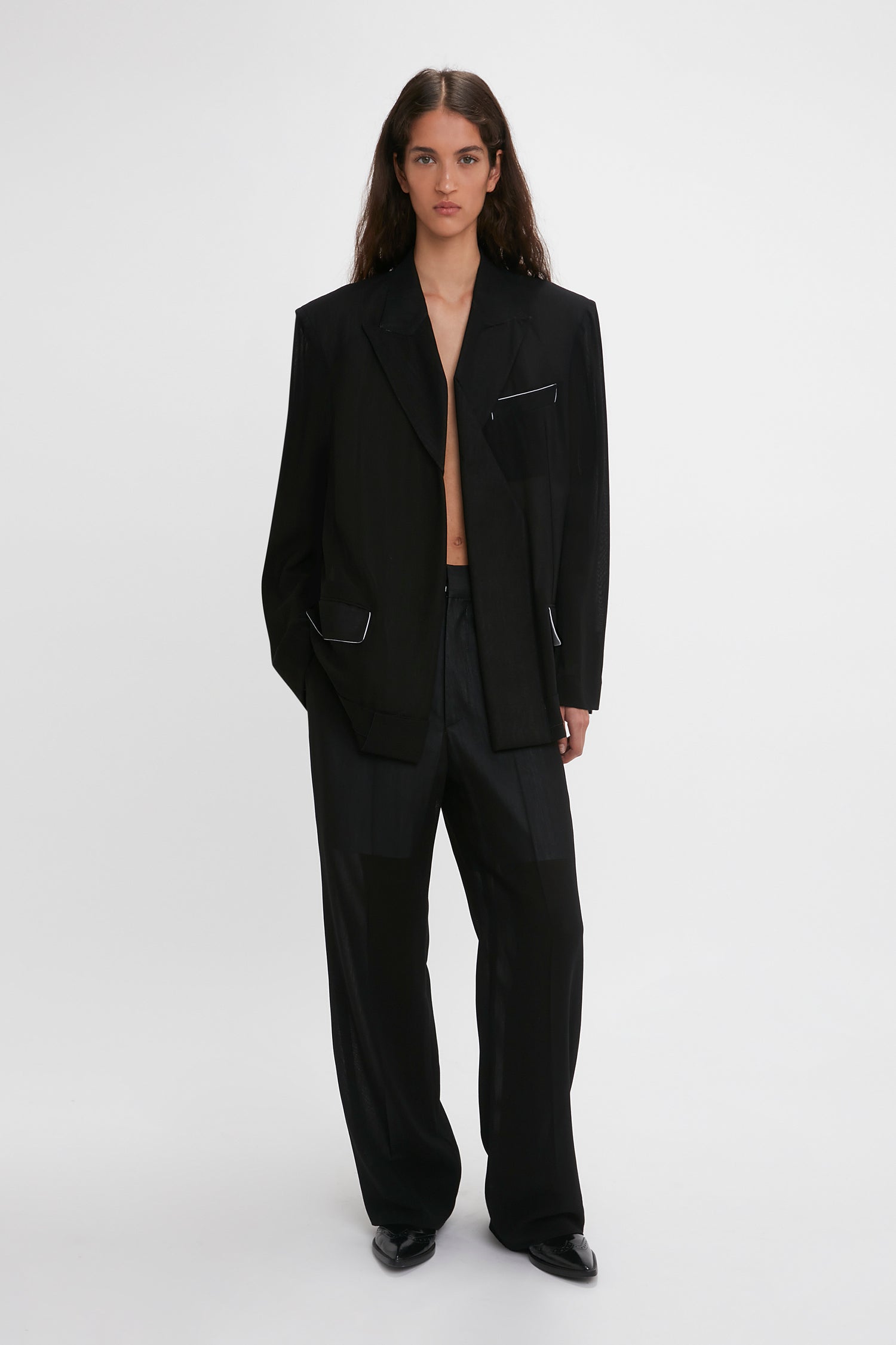 A woman models a Victoria Beckham black suit featuring a Fold Detail Tailored Jacket and straight leg trousers with waistband detail, standing against a plain white background.