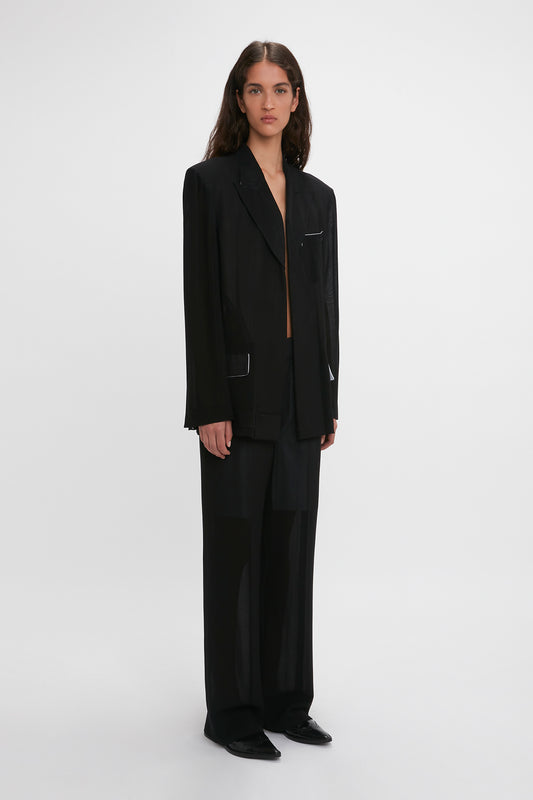 A woman stands facing the camera, wearing a sleek black pantsuit with a Victoria Beckham Fold Detail Tailored Jacket in Black and straight-leg trousers, on a plain white background.