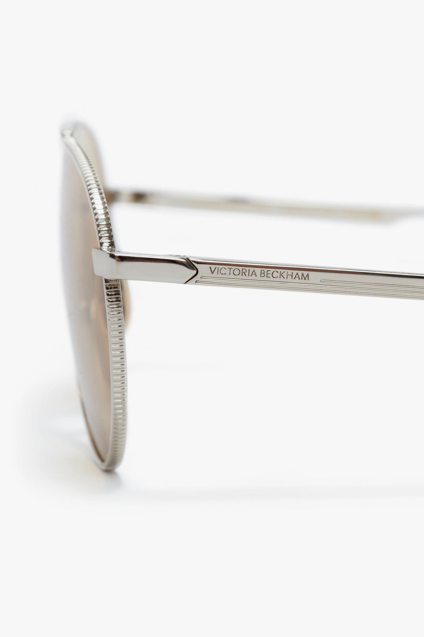 Close-up view of the side of a pair of Italian crafted Victoria Beckham V Metal Pilot Sunglasses In Silver-Brown, showing the brand name engraved on the temple, against a white background.