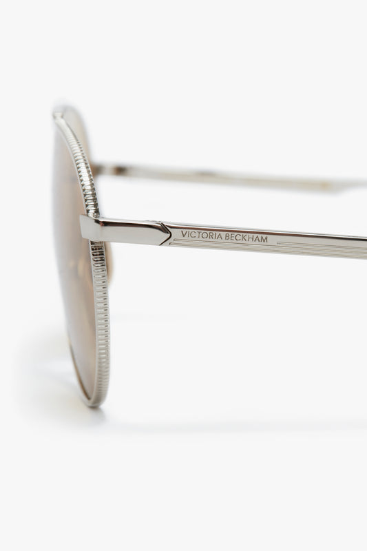 Close-up view of the side of a pair of Italian crafted Victoria Beckham V Metal Pilot Sunglasses In Silver-Brown, showing the brand name engraved on the temple, against a white background.