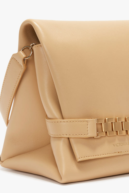 Beige leather crossbody bag with a fold-over flap, featuring a gold chain detail on the front. 

Chain Pouch With Strap In Sesame Leather by Victoria Beckham.