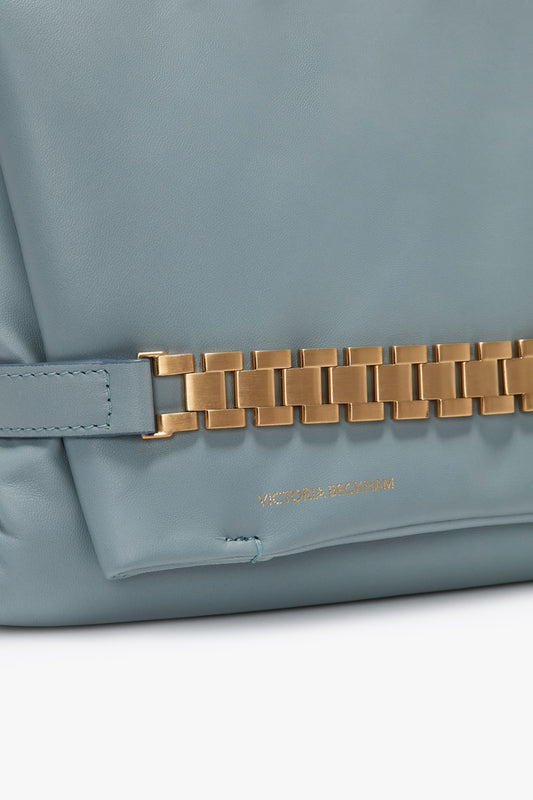 Close-up of a light blue Victoria Beckham Puffy Chain Pouch With Strap In Ice Leather handbag with a gold chain detail and "victoria beckham" embossed on the nappa leather.