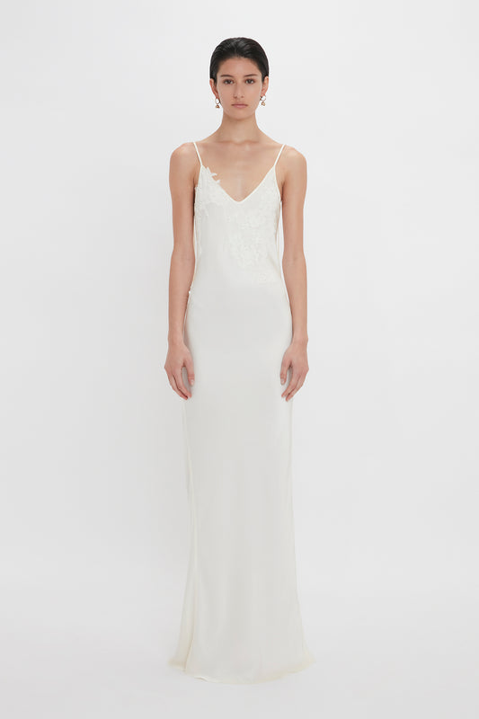 A woman in an elegant white sleeveless wedding dress with lace appliqué on the bodice, standing against a plain white background by Victoria Beckham.