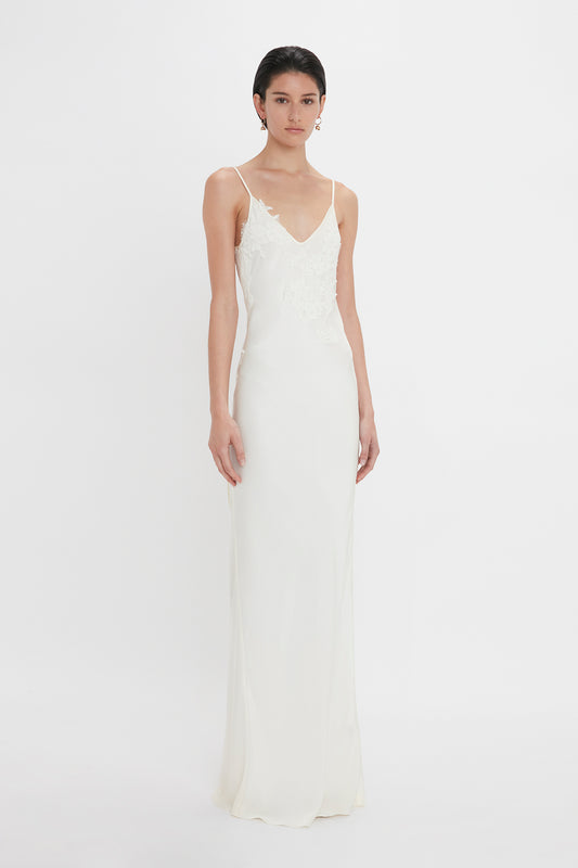 A woman in a sleek, white sleeveless wedding dress with lace detailing at the bodice, standing against a plain white background, wearing Victoria Beckham's Exclusive Camellia Flower Hoop Earrings In Gold.