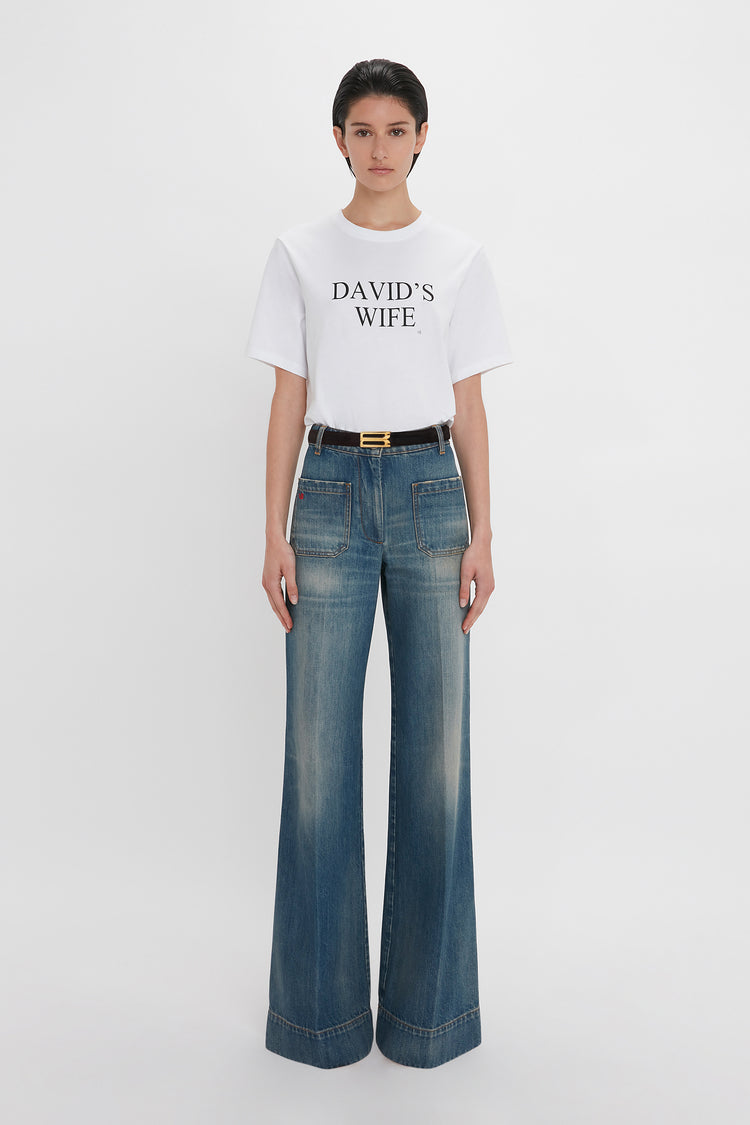 A woman in a Victoria Beckham 'David's Wife' slogan T-shirt in white and blue high-waisted jeans with a gold buckle belt, standing against a plain white background.