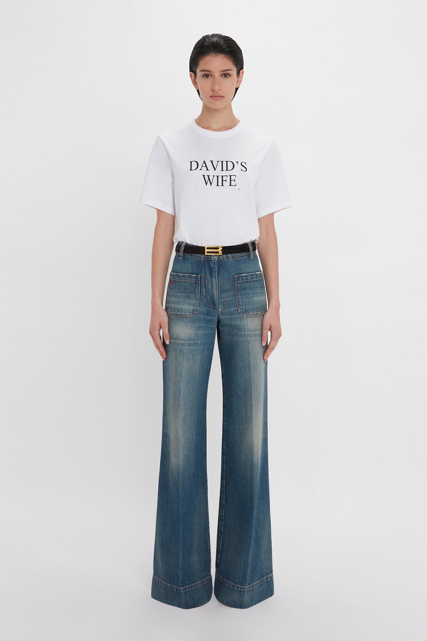 A woman in a Victoria Beckham 'David's Wife' slogan T-shirt in white and blue high-waisted jeans with a gold buckle belt, standing against a plain white background.