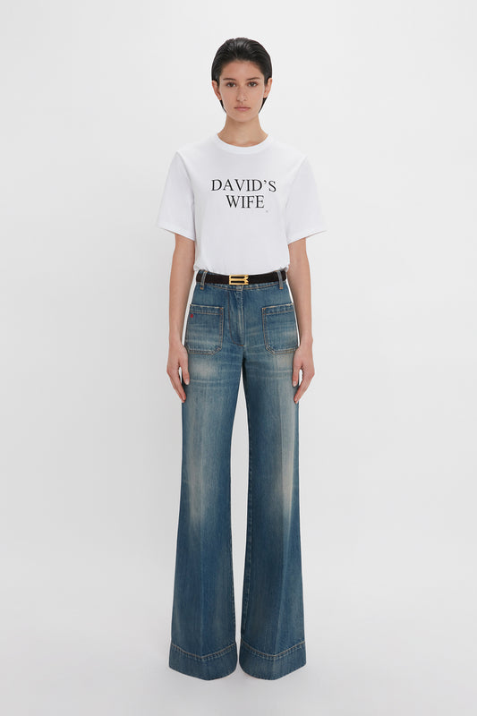 A woman in a Victoria Beckham 'David's Wife' slogan T-shirt in white and blue high-waisted jeans with a gold buckle belt, standing against a plain white background.