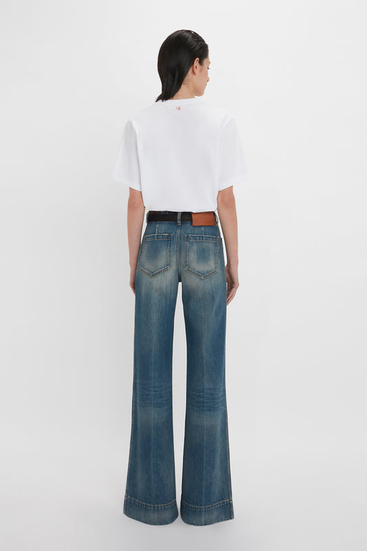 Rear view of a woman dressed in a white t-shirt and high-waisted flared Victoria Beckham Alina Jean In Indigrey Wash, standing against a plain white background.