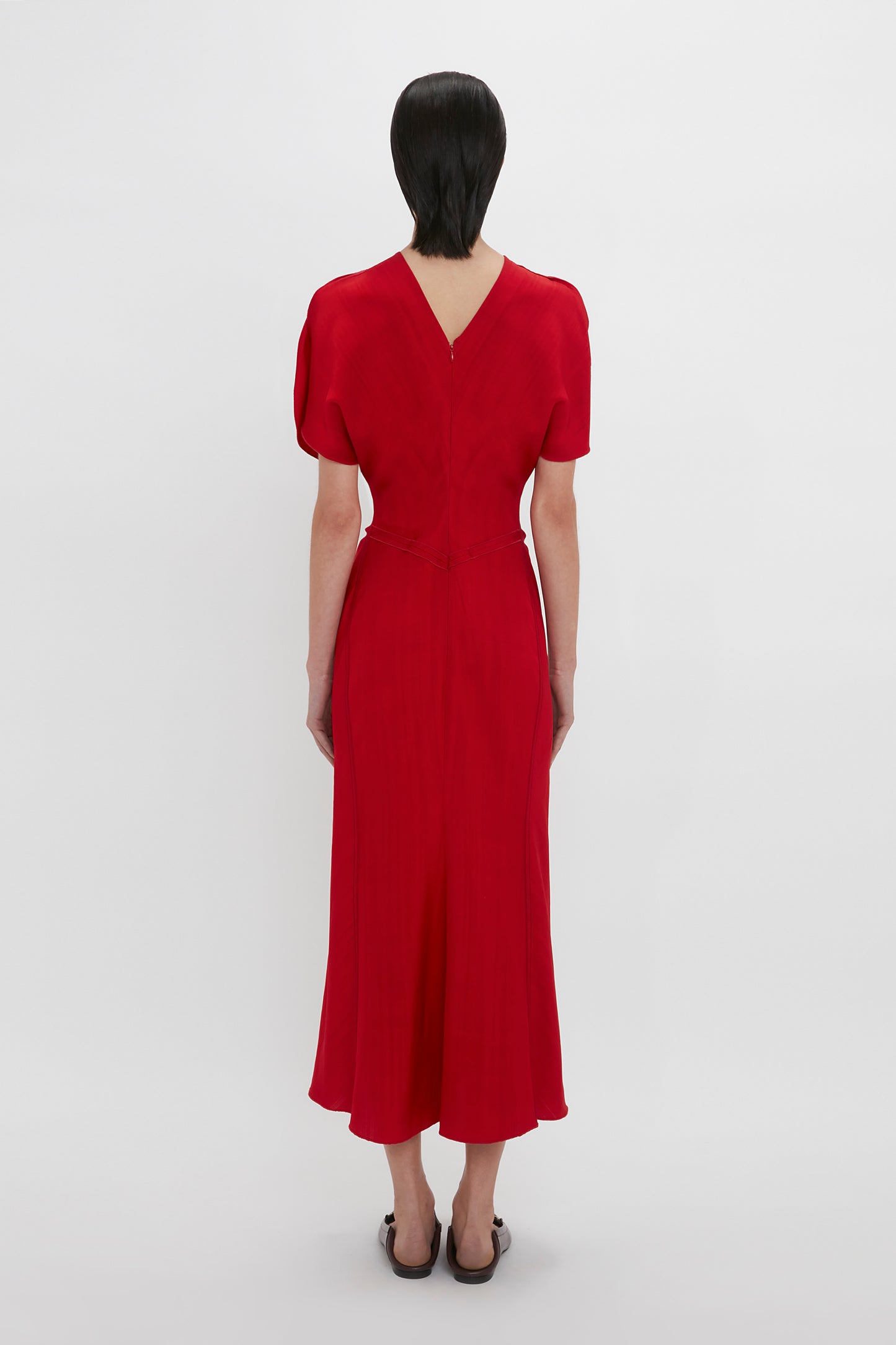 A woman stands facing away, wearing a Victoria Beckham Exclusive Gathered V-Neck Midi Dress In Carmine with short sleeves and a v-neckline against a plain white background.