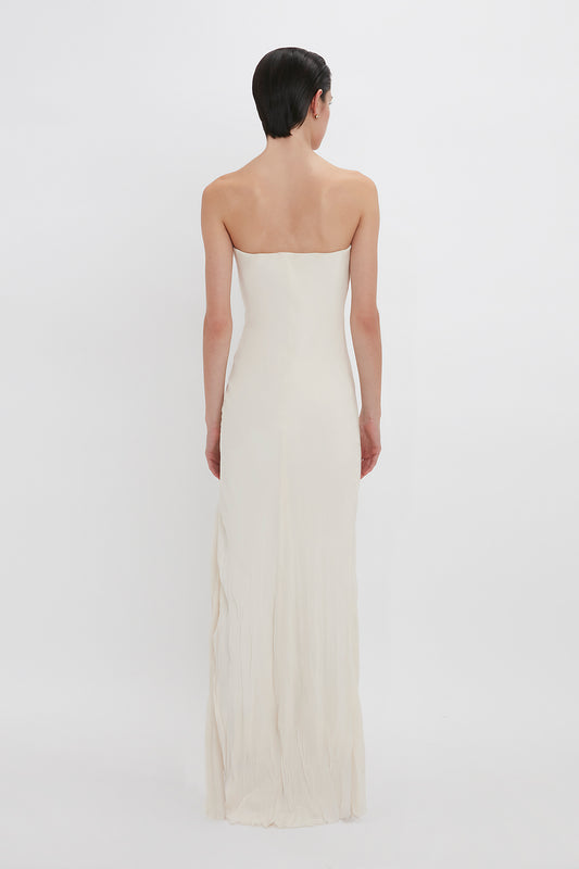 A woman with short black hair stands facing away, wearing a long, strapless sweetheart neckline, Victoria Beckham Exclusive Floor-Length Corset Detail Gown In Ivory against a plain white background.