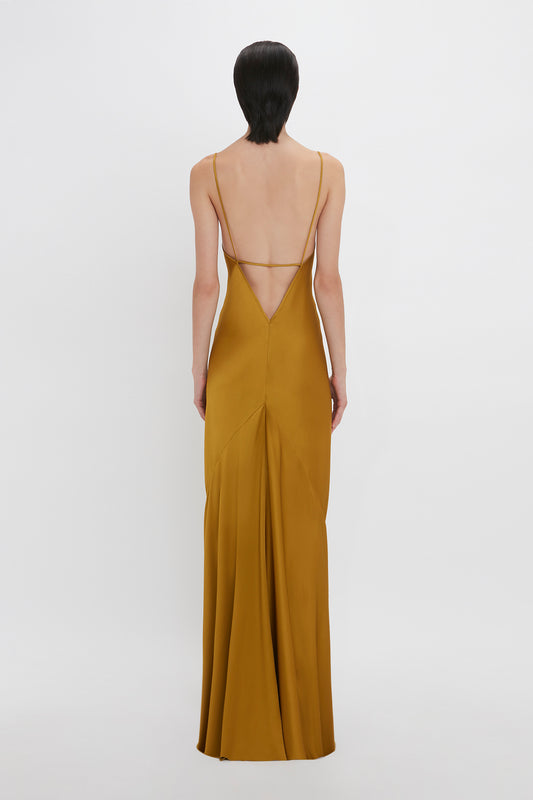 A woman standing with her back to the camera, wearing an elegant Victoria Beckham Low Back Cami Floor-Length Dress In Harvest Gold with a low-cut back and thin straps.