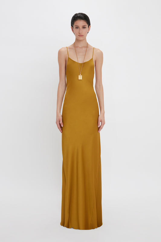 A woman in a sleek Victoria Beckham Low Back Cami Floor-Length Dress In Harvest Gold and a long necklace stands poised against a white background.