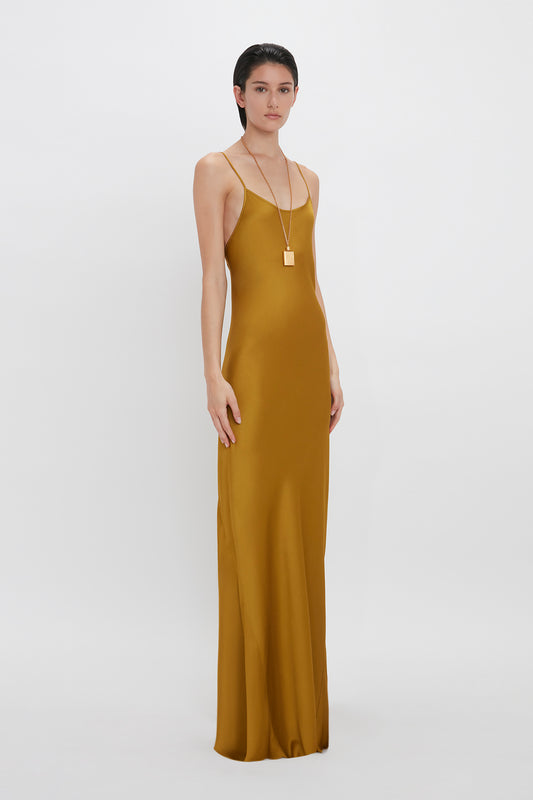 A woman in a sleek Victoria Beckham Low Back Cami Floor-Length Dress In Harvest Gold stands against a plain white background, accessorized with a long gold necklace.