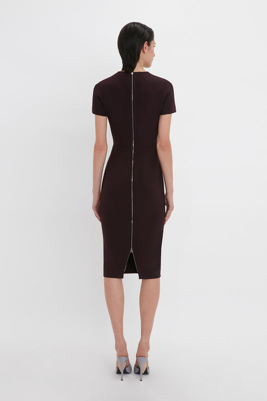 A woman in a Victoria Beckham fitted T-shirt dress in deep mahogany made of stretch double wool crepe and gray heels stands with her back to the camera against a white background.