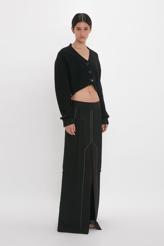 Woman standing in a studio, wearing a black Victoria Beckham cropped V-neck cardigan with detailed stitching and high-waisted pants.
