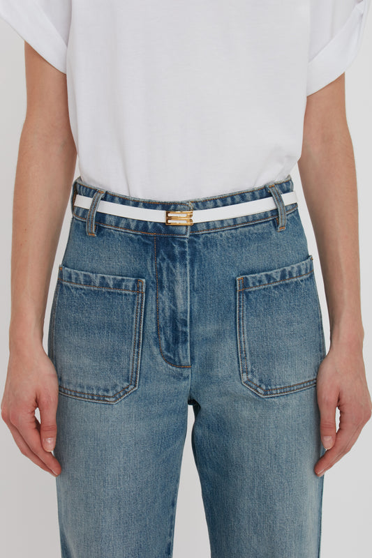 Close-up of a person wearing blue jeans and a white t-shirt, with hands gently resting by their sides, focusing on a Victoria Beckham Exclusive Micro Frame Belt in White Leather with gold hardware.