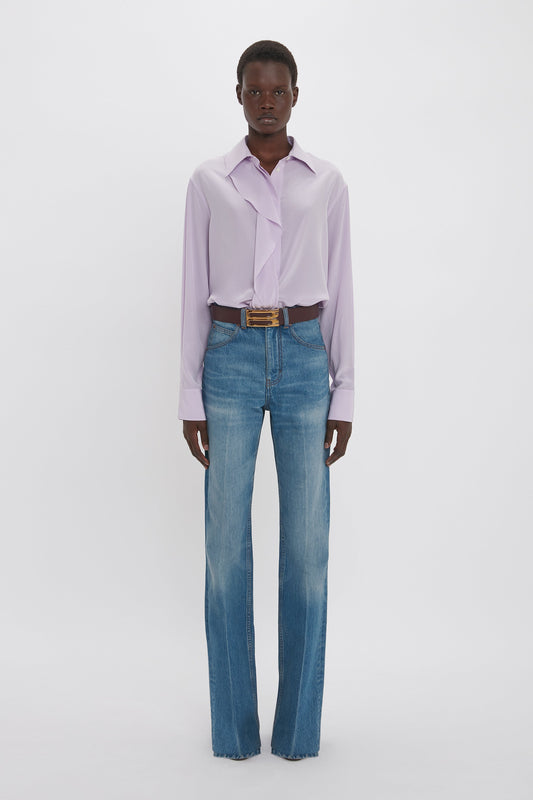 A black woman stands against a white background, wearing a Victoria Beckham Asymmetric Ruffle Blouse In Petunia, blue jeans, and a brown belt.