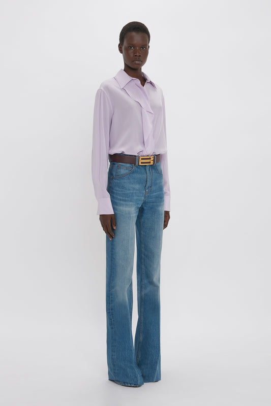 A young black man posing in a studio, wearing a Victoria Beckham Asymmetric Ruffle Blouse In Petunia, blue jeans, and a gold belt against a white backdrop.
