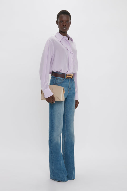 A woman stands wearing a Victoria Beckham Asymmetric Ruffle Blouse In Petunia, blue jeans, a gold belt, and holding a beige purse, against a white background.