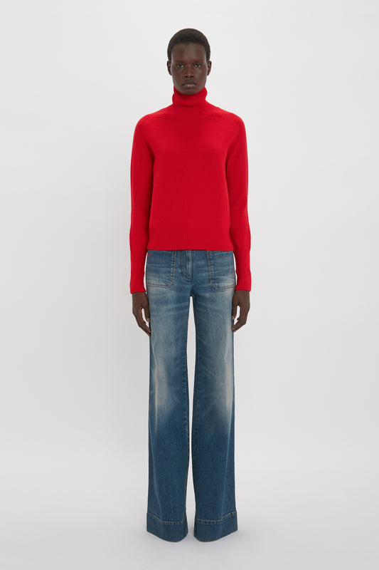 A man wearing a Victoria Beckham red lambswool polo neck jumper and blue jeans standing against a plain white background.