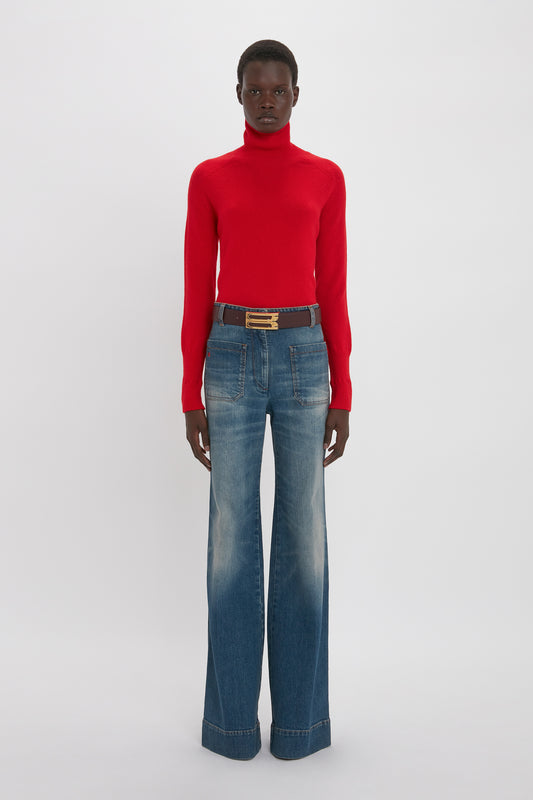 A young black woman in a fitted Victoria Beckham lambswool polo neck jumper in red and blue flared jeans, accessorized with a brown belt. She stands against a plain white background.