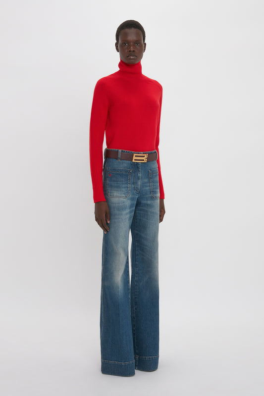 A person standing against a white background, wearing a Victoria Beckham red lambswool polo neck jumper and blue jeans with a gold-buckled belt.