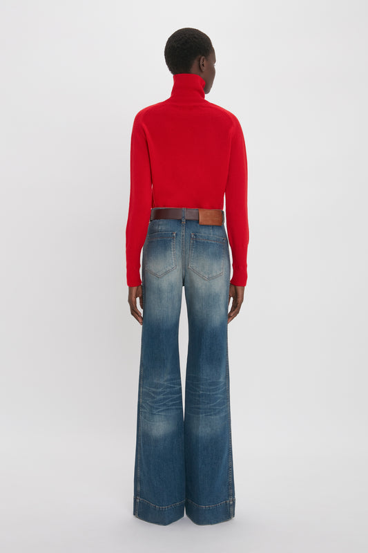 Rear view of a woman wearing a Victoria Beckham red lambswool polo neck jumper and blue wide-leg jeans, standing against a white background.