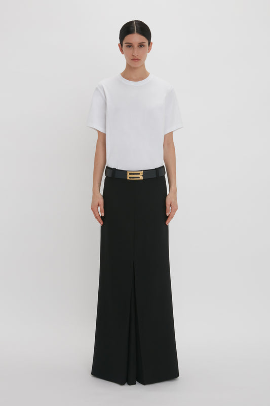 A woman in a white Victoria Beckham T-Shirt and black wide-legged trousers, wearing a black belt with a gold buckle, standing against a plain white background.