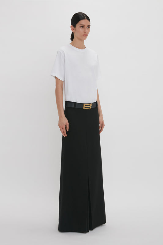 A woman in a Victoria Beckham luxury T-shirt and black wide-leg trousers with a gold buckle belt, standing against a plain white background.