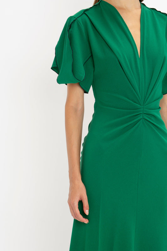 A close-up of a woman wearing a green Victoria Beckham Gathered V-Neck Midi Dress in Emerald with puffed sleeves and a twisted detail at the chest, on a white background.