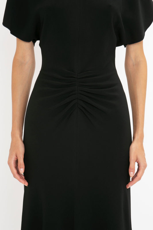 Close-up of a woman in a Victoria Beckham black fit-and-flare silhouette dress with flutter sleeves and a gathered waist detail, standing against a white background.